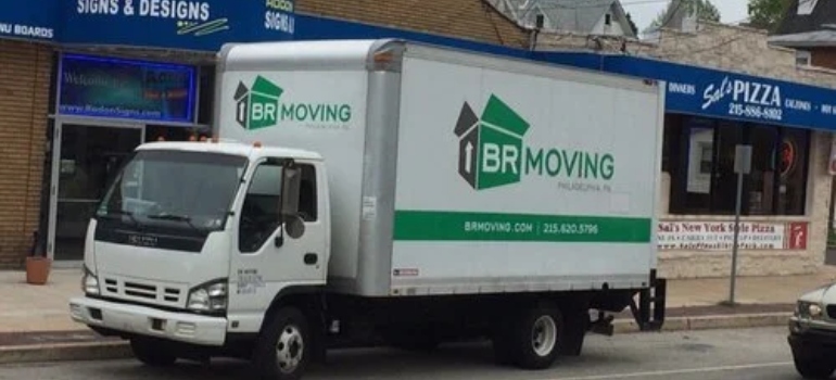 A white moving truck with "BR MOVING" logo parked on a street in front of a sign shop and a pizza restaurant.