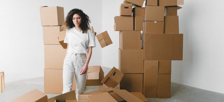 A young woman stands indoors surrounded by stacked cardboard boxes.